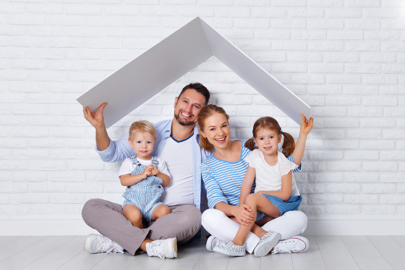 A family of four sitting against a white brick wall holding a white roof structure above their heads