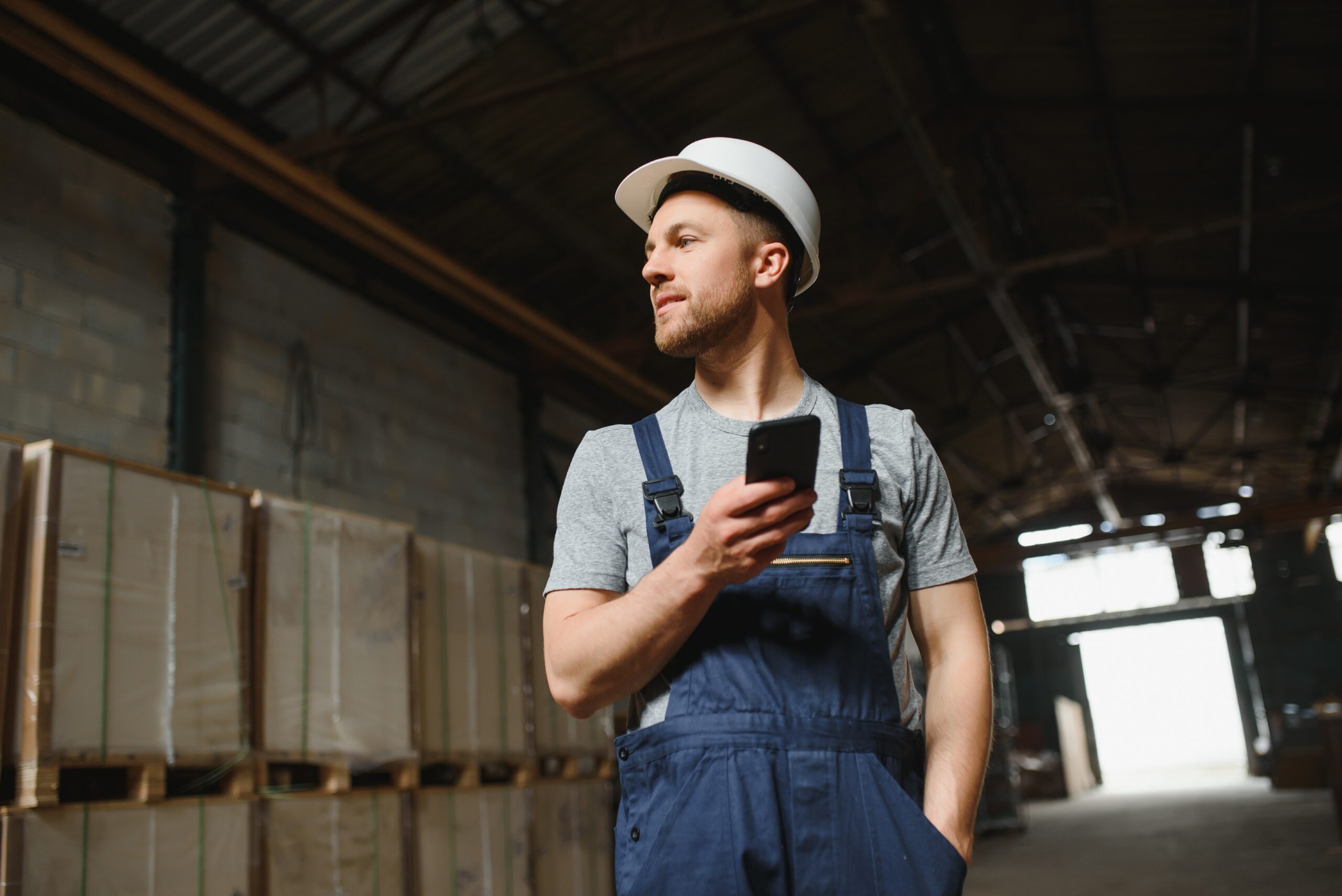 A man wearing work gear including hard hat is standing in an industrial building and holding his phone